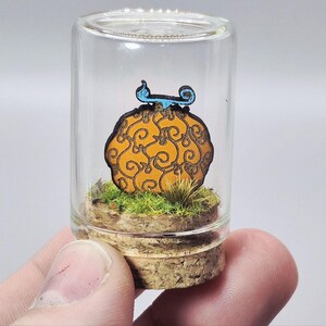 May include: A miniature, handcrafted wooden fruit with an orange and brown design, topped with a blue accent, encased in a clear glass jar. The fruit rests on a bed of green grass and soil, with a cork base.