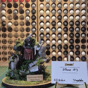 May include: A detailed figurine of Alphonse Elric from Fullmetal Alchemist, with a certificate of authenticity. The figure is gray and red, set on a round base with green moss. The background shows paint bottles.