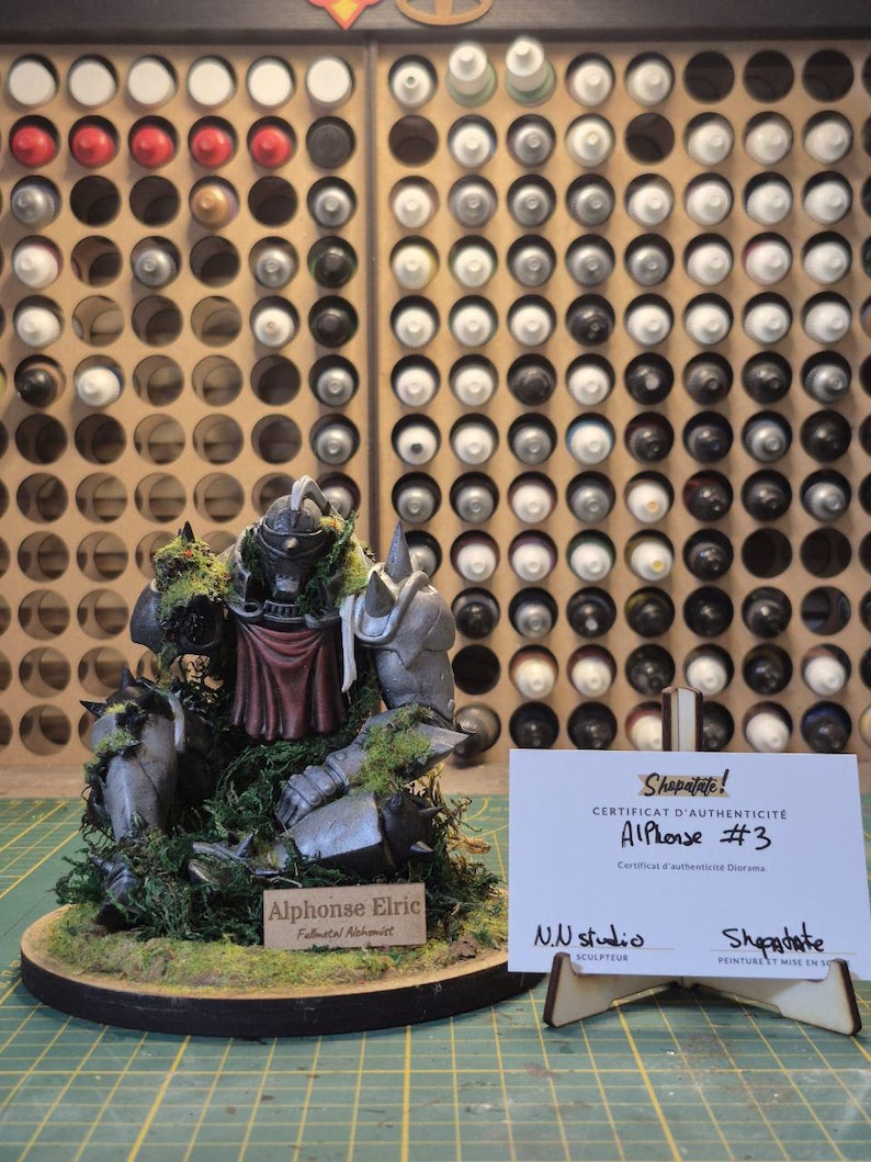 May include: A detailed figurine of Alphonse Elric from Fullmetal Alchemist, with a certificate of authenticity. The figure is gray and red, set on a round base with green moss. The background shows paint bottles.