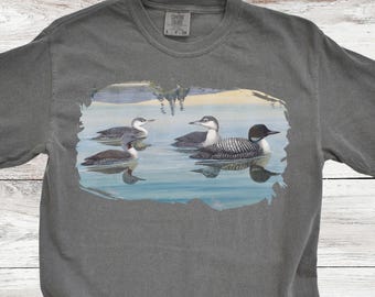 May include: A gray t-shirt with a print of three loons swimming in a lake. The loons are black and white with red eyes. The lake is blue and green. The text "Pepper" is in the upper right corner of the image.