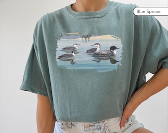 May include: A teal blue t-shirt with a graphic print of four common loons swimming in a lake. The text "Blue Spruce" is in the top right corner of the image.