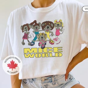 Puede incluir: Camiseta blanca con un gráfico de cinco ratones de dibujos animados con atuendos coloridos y el texto "Mice World". La camiseta también tiene una pequeña bandera canadiense con el texto "Designed & Printed in Canada".