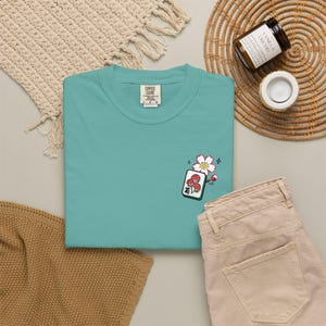 May include: A teal t-shirt featuring a small embroidered design on the chest. The embroidery depicts a white cherry blossom flower positioned above a rectangular mahjong tile that displays a red floral pattern and black Chinese characters.