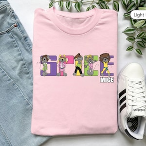 Puede incluir: Camiseta rosa claro con la frase "SPACE MICE" en letras coloridas. Cada letra presenta un personaje de ratón de dibujos animados con diferentes atuendos. La camiseta está junto a zapatillas blancas y unos vaqueros azules.