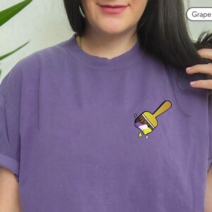 May include: Purple t-shirt with a yellow paintbrush design. The paintbrush is dripping with purple, yellow, and white paint.