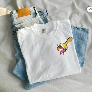 May include: White t-shirt with a paintbrush design featuring the colors of the lesbian pride flag.