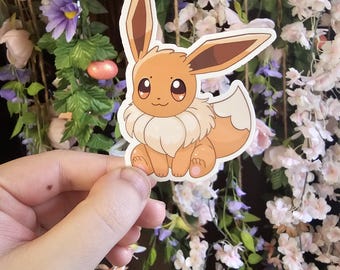 May include: A brown and tan cartoon Eevee Pokemon sticker with big eyes and a fluffy tail. The sticker is held in a hand against a background of pink and white flowers.