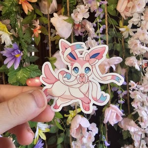 以下が含まれることがあります： ピンクとブルーの漫画のステッカー。ポケモンのキャラクター、シルヴァディ。ピンクのリボンをつけています。ステッカーは、ピンクと白の花の背景の前にある手のひらの上にあります。