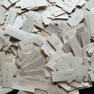May include: A pile of vintage flashcards with printed words and phrases. The cards are off-white and rectangular, with black text. The cards are scattered, creating a textured, layered effect. Some cards are stacked, while others are face up.