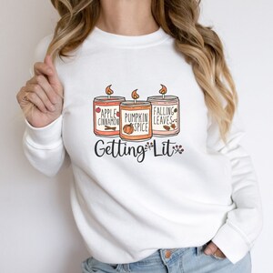 May include: White sweatshirt with a graphic of three candles with fall scents: apple cinnamon, pumpkin spice, and falling leaves. The text "Getting Lit" is below the candles.