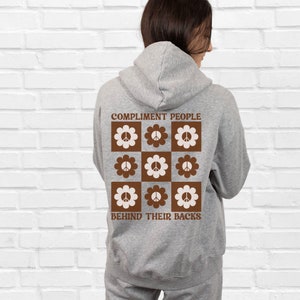 May include: A gray hoodie with a brown and white peace sign design on the back. The text "COMPLIMENT PEOPLE BEHIND THEIR BACKS" is printed on the hoodie.