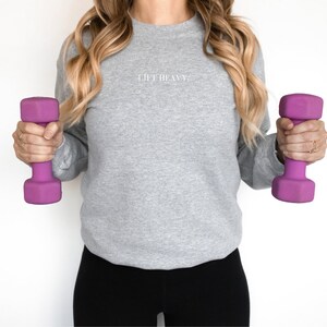 May include: A woman wearing a gray sweatshirt with the text "Lift Heavy" on the front, holding a purple dumbbell in each hand.