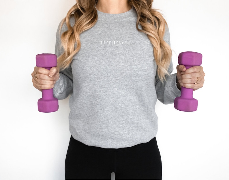 May include: A woman wearing a gray sweatshirt with the text "Lift Heavy" on the front, holding a purple dumbbell in each hand.