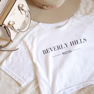 May include: A white t-shirt with the words "BEVERLY HILLS 90210" printed in black. A beige fedora hat and a cream-colored handbag with gold-tone hardware are also in the image. The items are arranged on a soft, textured surface.