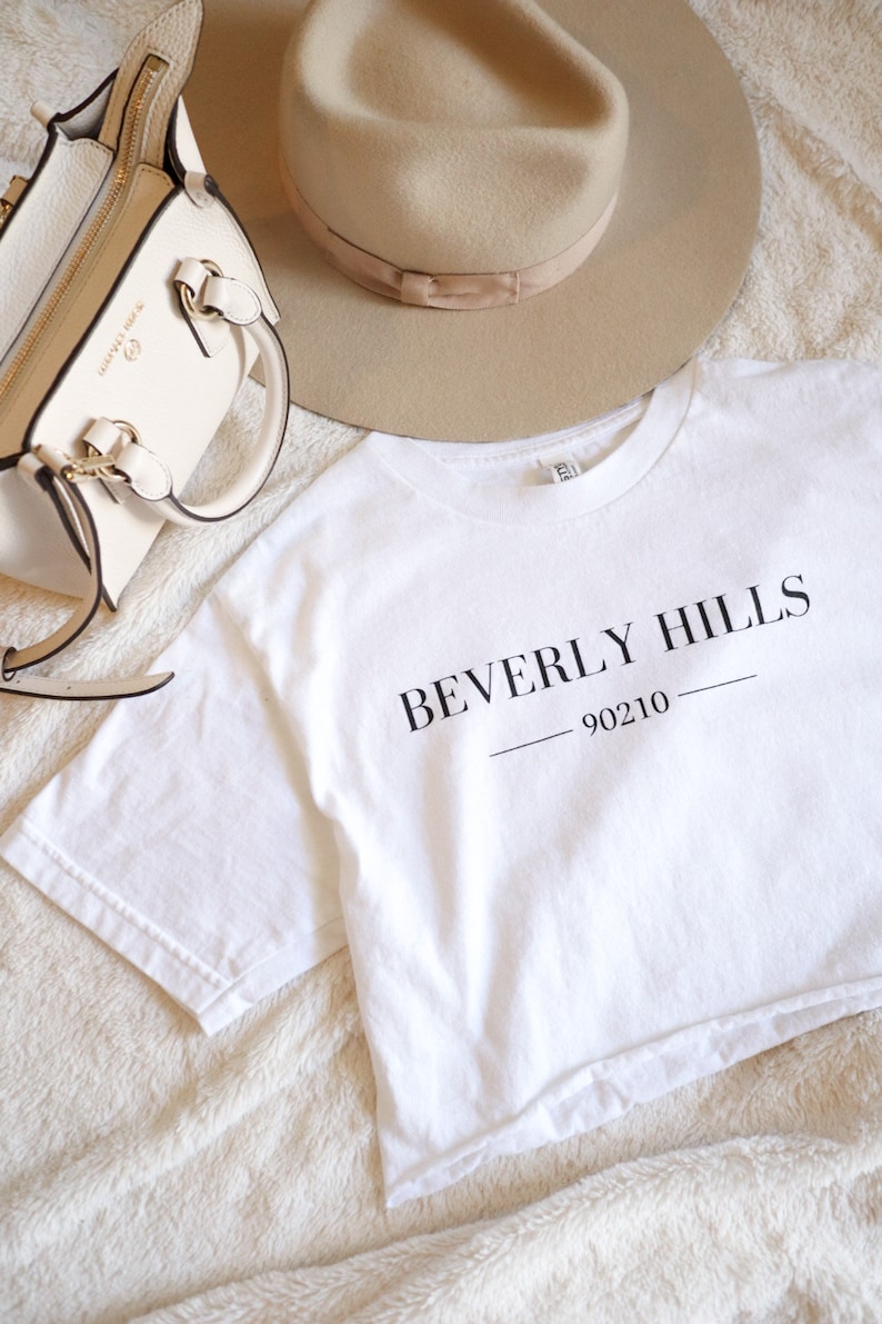 May include: A white t-shirt with the words "BEVERLY HILLS 90210" printed in black. A beige fedora hat and a cream-colored handbag with gold-tone hardware are also in the image. The items are arranged on a soft, textured surface.