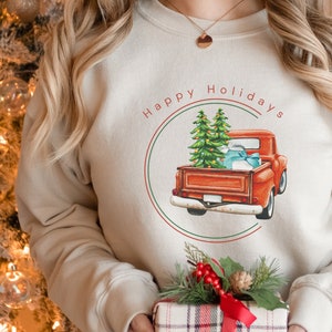 May include: A beige sweatshirt with a red truck carrying Christmas trees and presents. The truck is inside a circle with the text "Happy Holidays" in red and green.