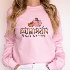 May include: A light pink sweatshirt featuring the text "autumn wishes PUMPKIN kisses" with pumpkin graphics. The pumpkins are orange and pink with heart patterns. The sweatshirt is a long-sleeved crew neck.