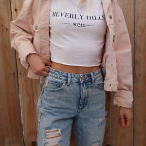 May include: A light pink corduroy jacket worn over a white crop top with "BEVERLY HILLS 90210" printed in black. The outfit includes light blue ripped jeans. The background is a wooden fence.