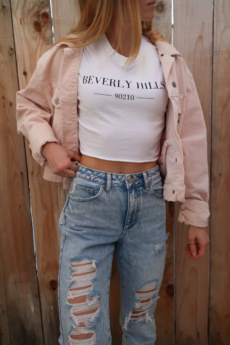 May include: A light pink corduroy jacket worn over a white crop top with "BEVERLY HILLS 90210" printed in black. The outfit includes light blue ripped jeans. The background is a wooden fence.
