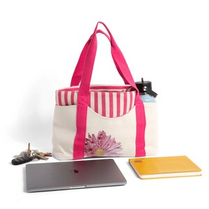 May include: A white canvas tote bag with pink handles and a pink and white striped lining. The bag has a large pink daisy embroidered on the front. The bag is filled with a laptop, a notebook, and a water bottle.