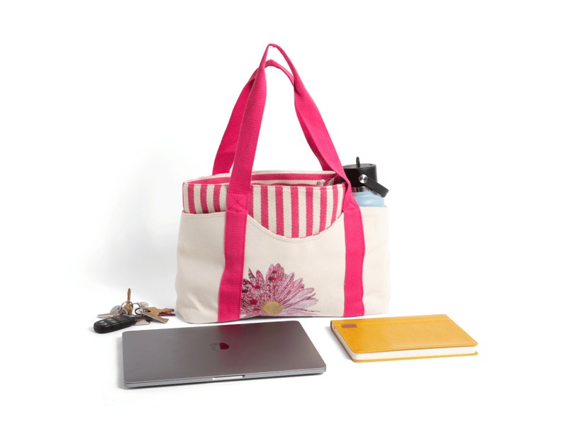 May include: A white canvas tote bag with pink handles and a pink and white striped lining. The bag has a large pink daisy embroidered on the front. The bag is filled with a laptop, a notebook, and a water bottle.