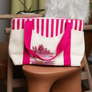 May include: A white canvas tote bag with pink stripes and a pink flower design. The bag has two pink handles.