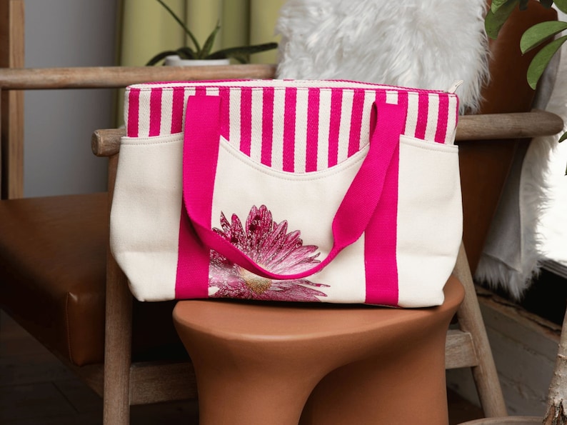 May include: A white canvas tote bag with pink stripes and a pink flower design. The bag has two pink handles.