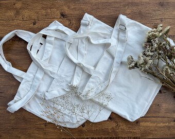 May include: Three white canvas tote bags with handles. The bags are folded and stacked on top of each other. Dried white flowers are scattered around the bags.