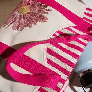 May include: A white canvas tote bag with pink stripes and a pink floral design. The bag has two pink handles and a blue water bottle is partially visible inside the bag.