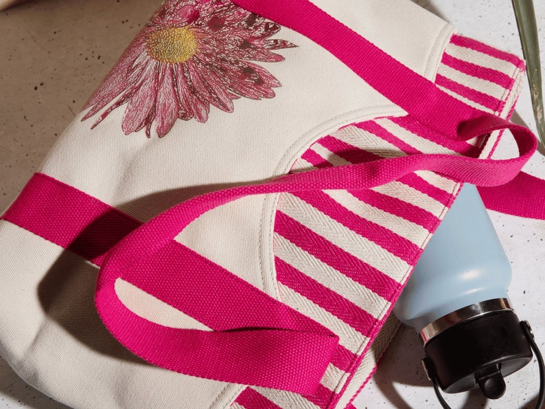 May include: A white canvas tote bag with pink stripes and a pink floral design. The bag has two pink handles and a blue water bottle is partially visible inside the bag.