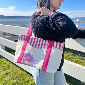 May include: A white canvas tote bag with pink and white stripes, a pink handle, and a pink flower design. The bag has a large pocket on the front.