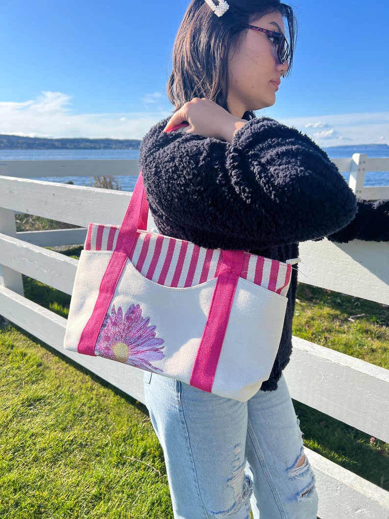 May include: A white canvas tote bag with pink and white stripes, a pink handle, and a pink flower design. The bag has a large pocket on the front.