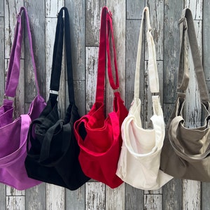May include: Five canvas tote bags hanging on a wooden wall. The bags are in different colors: purple, black, red, white, and gray.