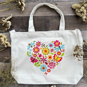 May include: White canvas tote bag with a colorful floral heart design. The heart is made up of flowers, butterflies, and dragonflies in shades of pink, red, yellow, blue, and green.