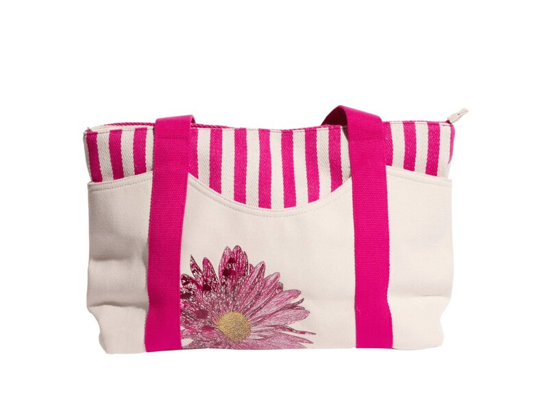 May include: A white canvas tote bag with pink stripes and a pink flower design. The bag has two pink handles.