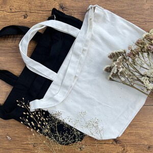 May include: Two tote bags, one black and one white, are lying on a wooden surface. Dried flowers are in the background.