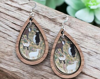May include: A pair of wooden teardrop-shaped earrings with a clear resin insert featuring a whimsical illustration of goats and flowers. The earrings have silver hooks.