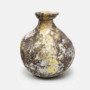 May include: A small, round ceramic vase with a narrow neck and a flared rim. The surface features a mottled, textured finish in shades of dark brown, charcoal gray, white, and pale yellow, creating a rugged, uneven appearance across the body.