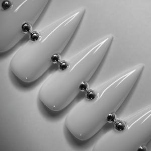 May include: A set of five glossy white press-on nails with a sharp stiletto shape. Each nail features two small, reflective silver metallic spheres placed horizontally across the center, creating a uniform and clean decorative accent on each piece.