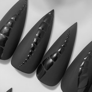 May include: A set of four matte black press-on nails with a sharp stiletto shape. Each nail features a raised, glossy black textured spine running down the center, resembling a reptile scale pattern that tapers toward the pointed tip of the nail.