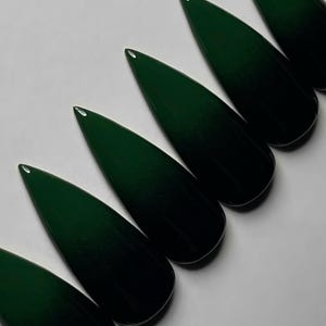 May include: A set of dark green, stiletto-shaped artificial nails. The nails are arranged diagonally across a white surface, showcasing their glossy finish and pointed tips. The color is a deep, rich green.