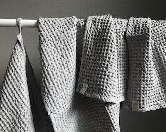 Puede incluir: Cuatro toallas de cocina de tejido de waffle gris colgadas de una barra blanca. Las toallas están hechas de un material suave y absorbente, perfectas para secar platos, manos y superficies.