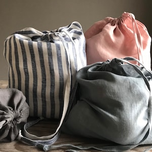 Può includere: Quattro sacchetti di lino con coulisse in diversi colori: grigio, blu e bianco a righe e rosa. I sacchetti sono realizzati in lino naturale e hanno un aspetto semplice e rustico.