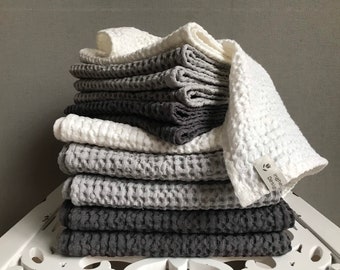 May include: A stack of waffle-weave towels in shades of white, gray, and charcoal gray. The towels are neatly folded and stacked on top of each other. One towel has a tag that reads "Ingrid Design".
