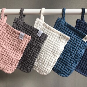 May include: Five kitchen pot holders hanging on a white rod. The pot holders are made of a textured fabric and come in five colors: pink, gray, white, blue, and dark blue.