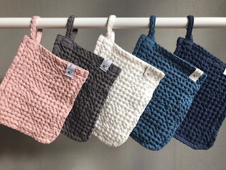May include: Five kitchen pot holders hanging on a white rod. The pot holders are made of a textured fabric and come in five colors: pink, gray, white, blue, and dark blue.