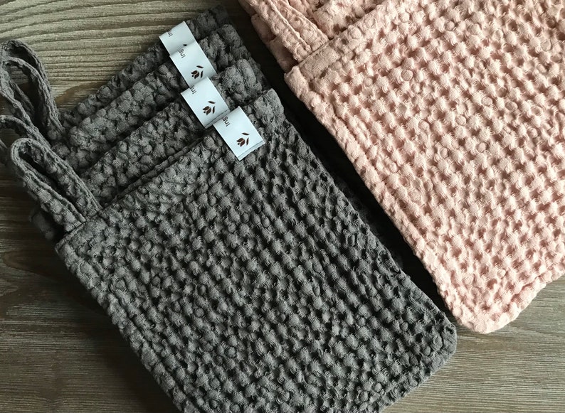 May include: Four gray waffle weave kitchen cloths with a hanging loop and a white label that says "leaf" stacked on top of each other. A pink waffle weave kitchen cloth is visible in the background.
