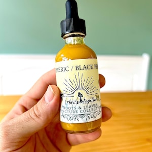 May include: A small glass bottle with a black dropper top, filled with a yellow liquid. The label reads "Turmeric / Black Pepper" and "Rhize Together Roots & Leaves Tincture Collection". The bottle is held in a hand.