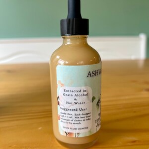 Op de afbeelding: Een heldere glazen fles met een zwarte druppelaar, gevuld met een lichtbruine vloeistof. Het etiket leest "ASHW" en "FOUR FLUID OUNCES". Het etiket bevat ook gebruiksaanwijzingen.