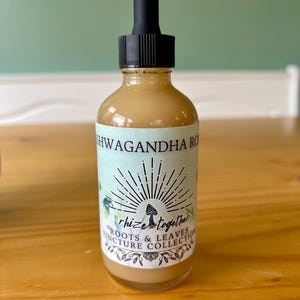 Op de afbeelding: Een heldere glazen fles met een zwarte druppelaar, gevuld met een lichtbruine vloeistof. Het etiket leest "Ashwagandha Roots & Leaves Tincture Collection" met een zonnestraalontwerp en het woord "rhize together". De fles staat op een houten oppervlak.